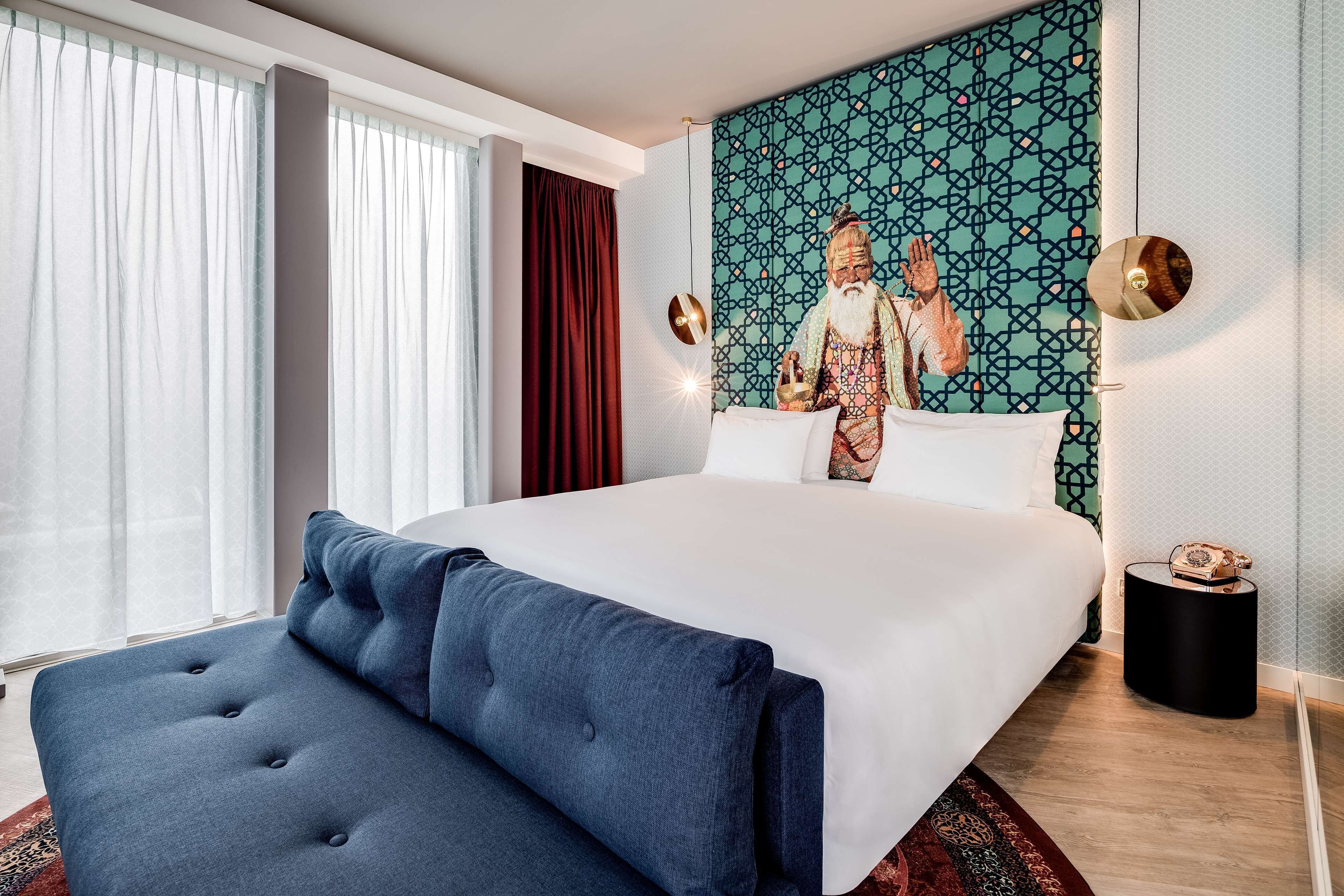 Une chambre d’hôtel moderne avec un grand lit, un banc recouvert d’un tissu bleu, une fresque murale à motifs et une petite table ronde avec un téléphone.