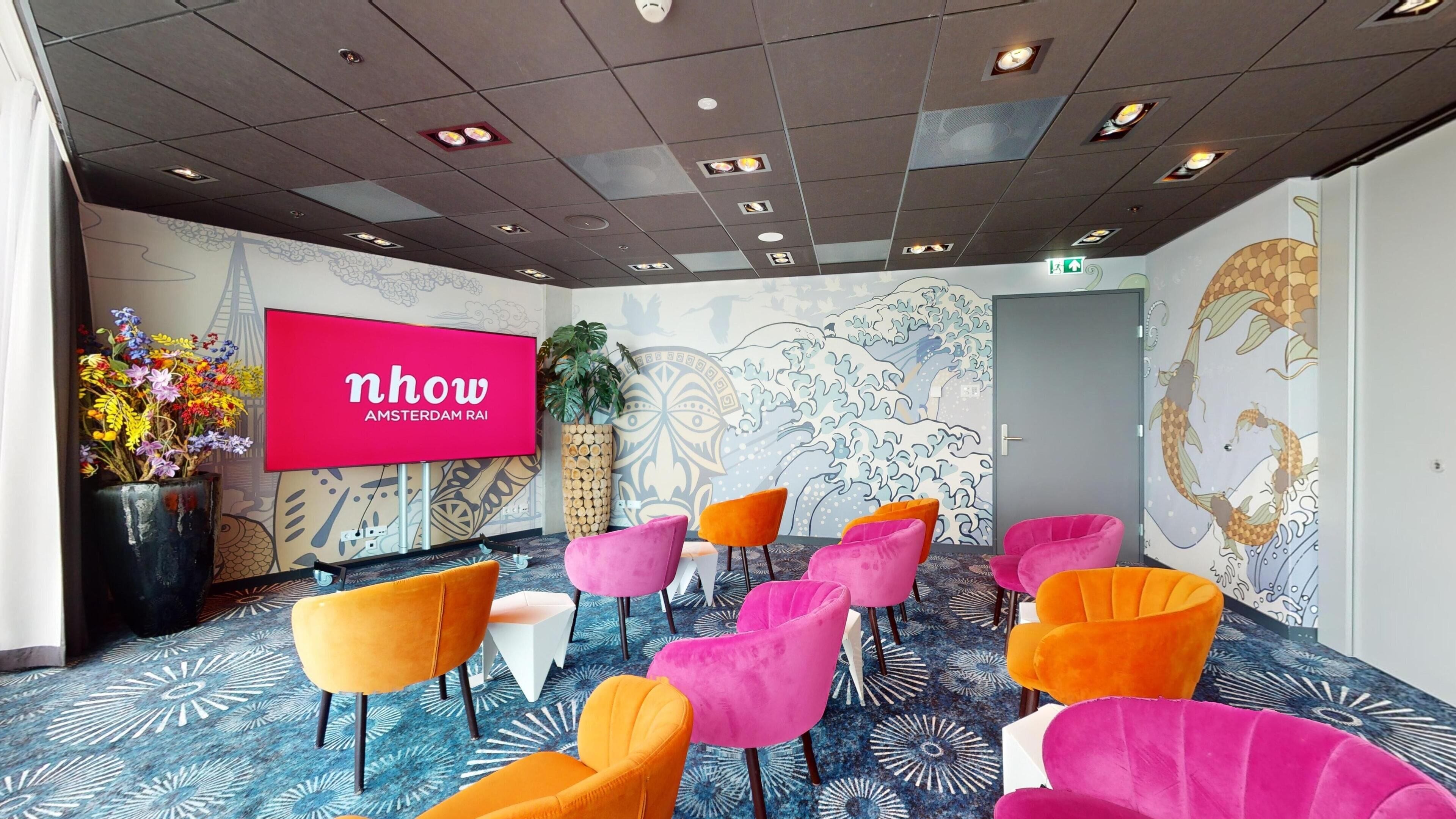 Ein Konferenzraum mit einer großen Leinwand, auf der 'nhow AMSTERDAM RAI' angezeigt wird, bunten Stühlen, einem gemusterten Teppich und einem Wandbild mit einem Fischmotiv.