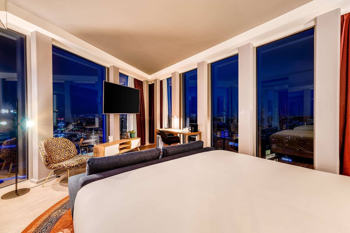 A modern hotel room with a large bed, a desk, a chair, and a TV.