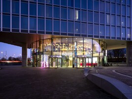 Un bâtiment moderne doté d’une façade en verre, d’une porte tournante à l’entrée et d’un intérieur bien éclairé.