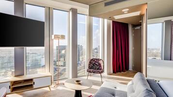A modern living room with large windows, a flat-screen TV, a sofa, a chair, and a small table.