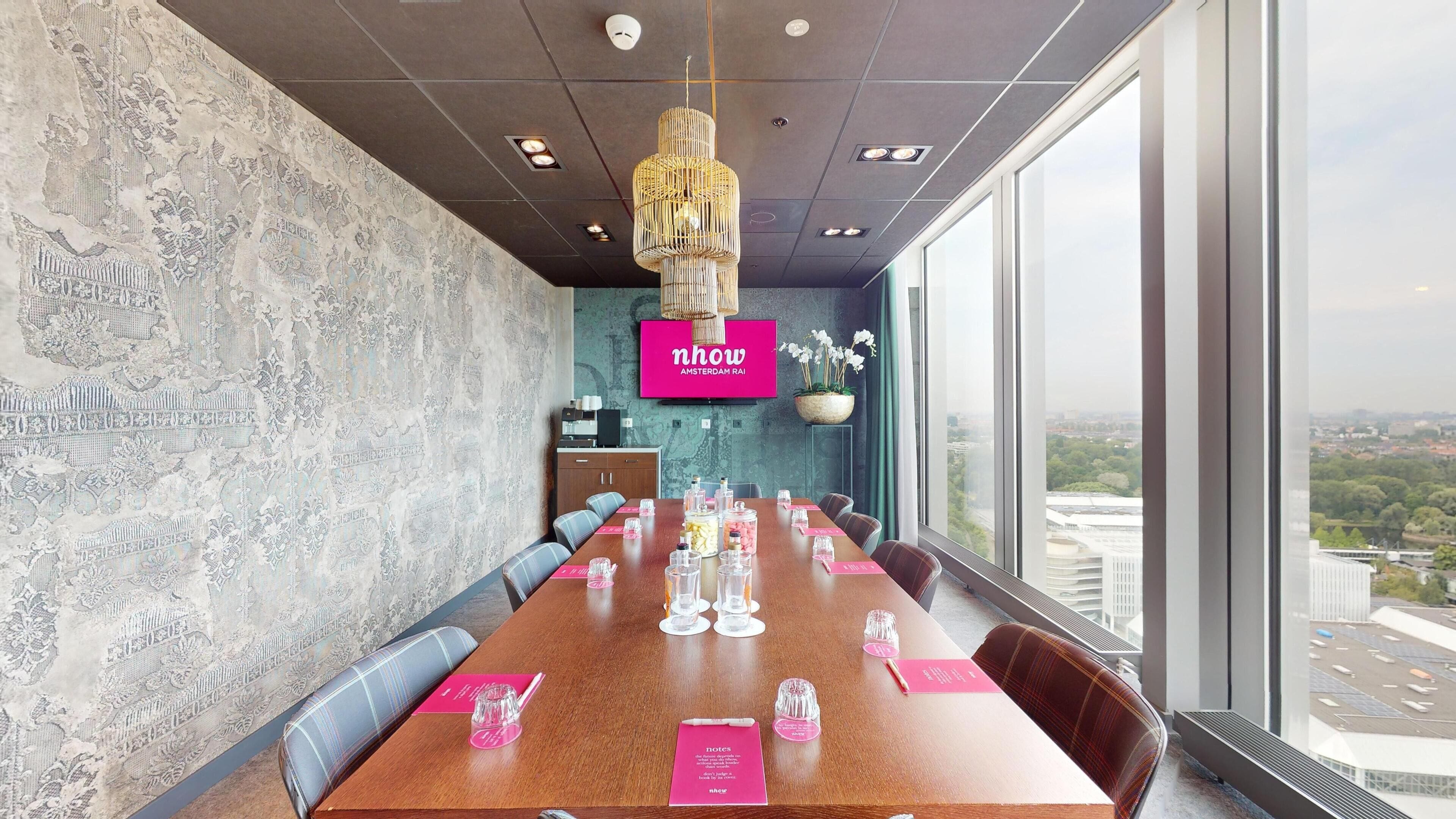 Een vergaderruimte met een lange tafel, stoelen, een groot raam met uitzicht op de stad, een decoratieve wand en een hanglamp.