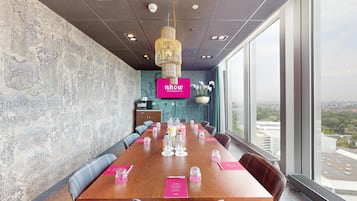 Een vergaderruimte met een lange tafel, stoelen, een groot raam met uitzicht op de stad, een decoratieve wand en een hanglamp.