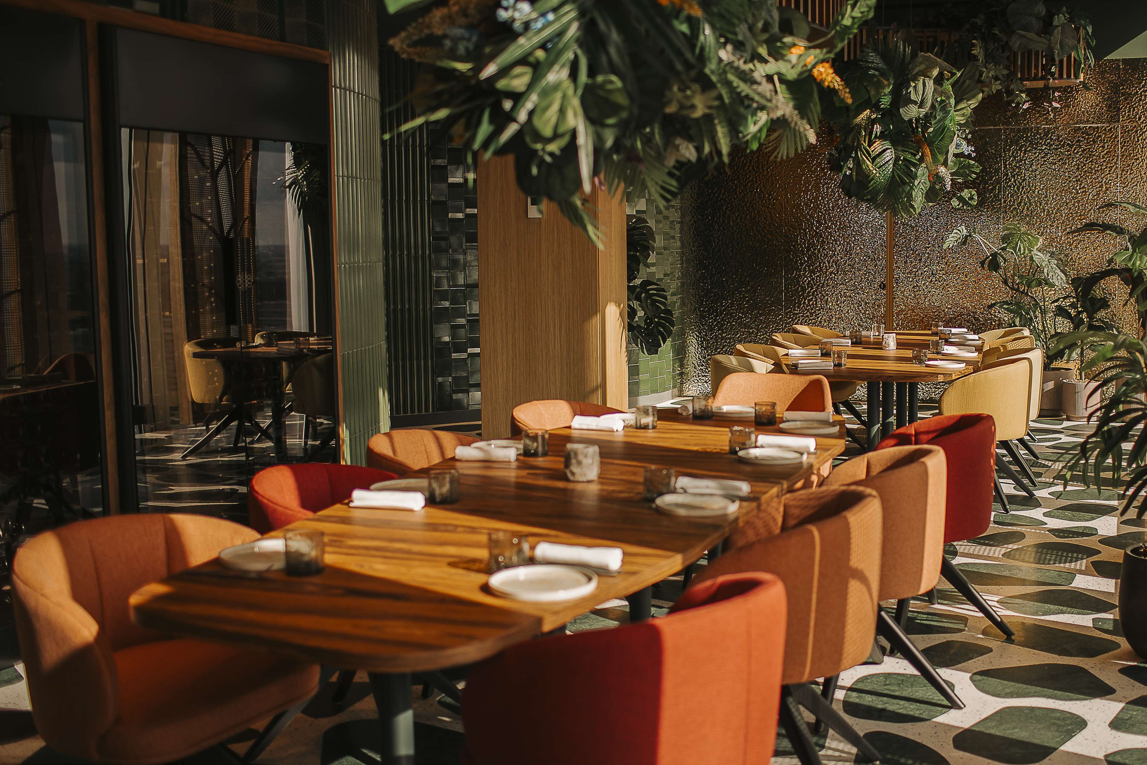 Un restaurante con mesas de madera y sillas naranjas, rodeado de plantas y una pared con textura.