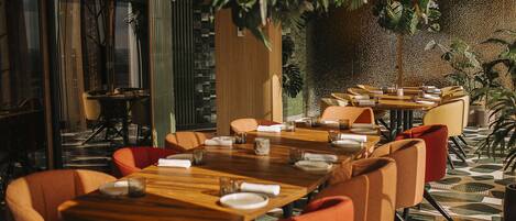 Un restaurante con mesas de madera y sillas naranjas, rodeado de plantas y una pared con textura.