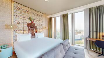 A hotel room with a large bed, a desk, a chair, and a view of the city through large windows.