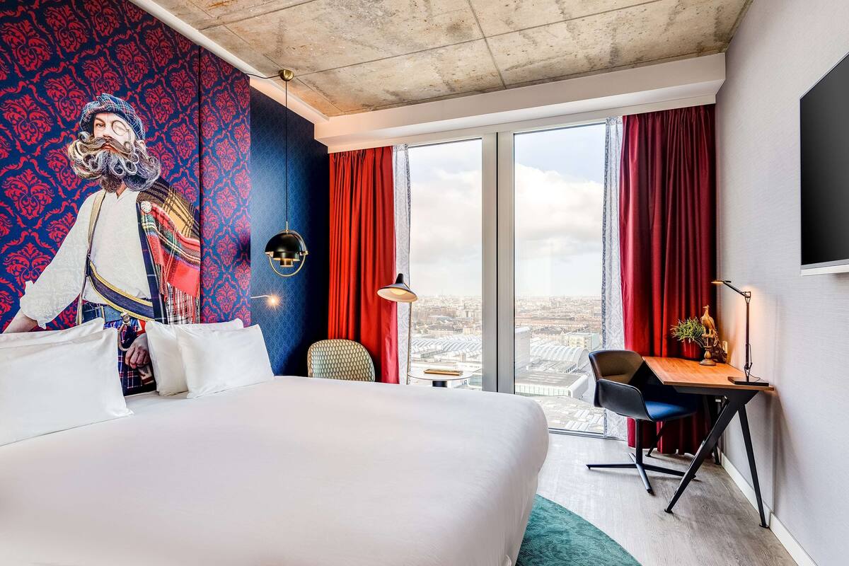 A hotel room with a large bed, a desk, a chair, a large window with red curtains, and a city view.