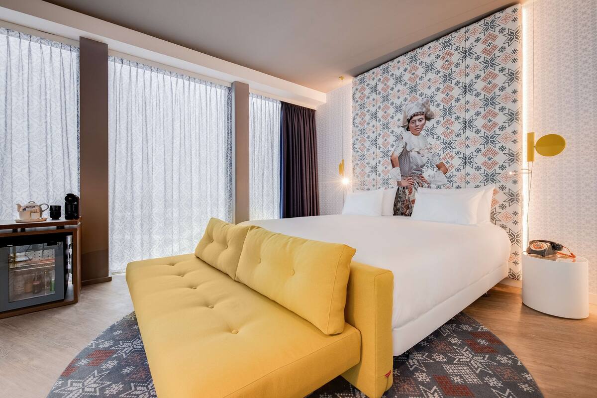 A modern hotel room with a large bed, a yellow sofa, a desk with a coffee machine, and patterned wallpaper.