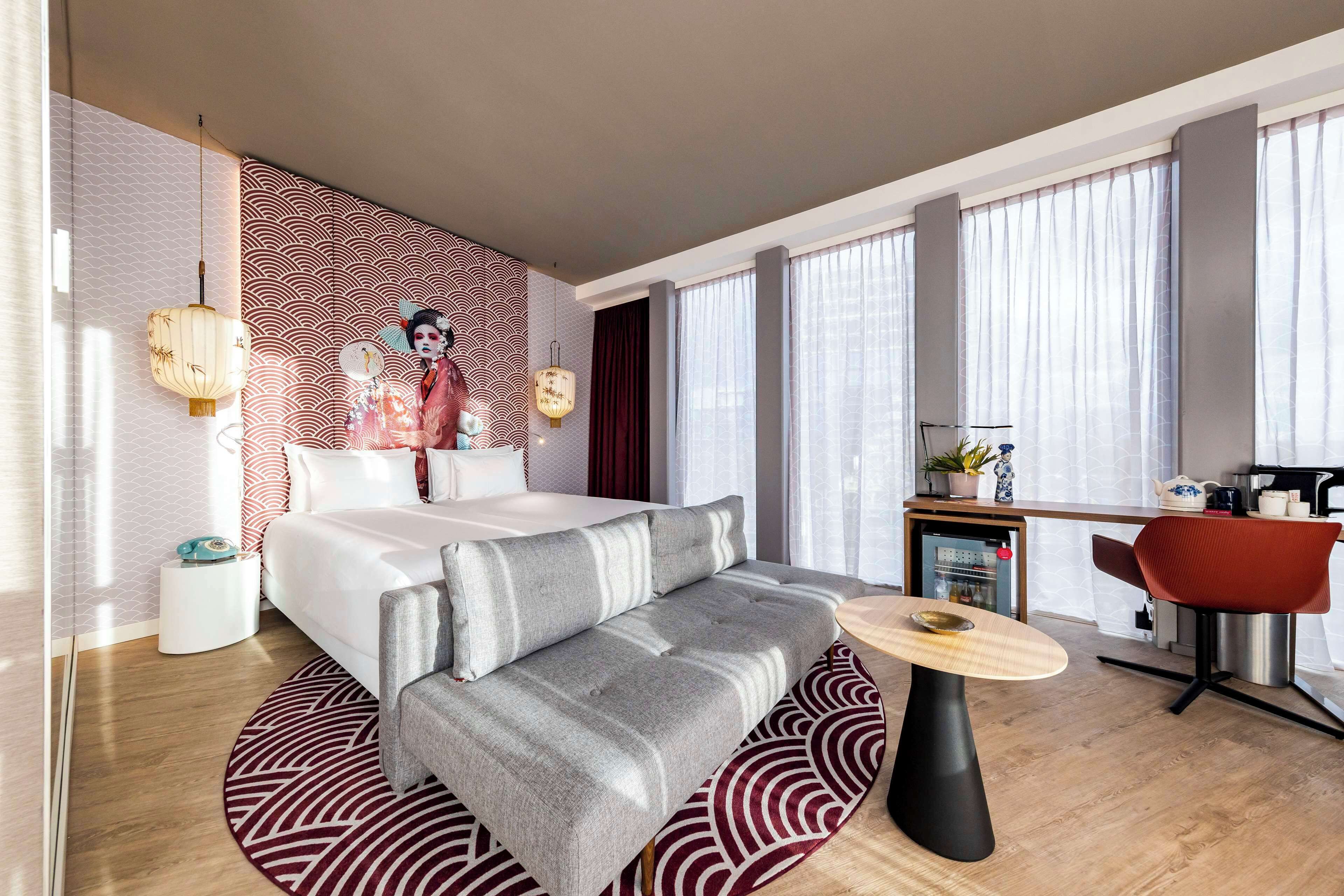 A modern hotel room with a bed, sofa, dining table, and kitchen area.