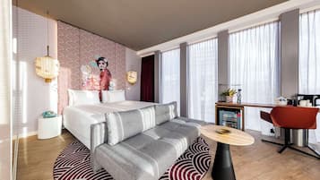 A modern hotel room with a bed, sofa, dining table, and kitchen area.
