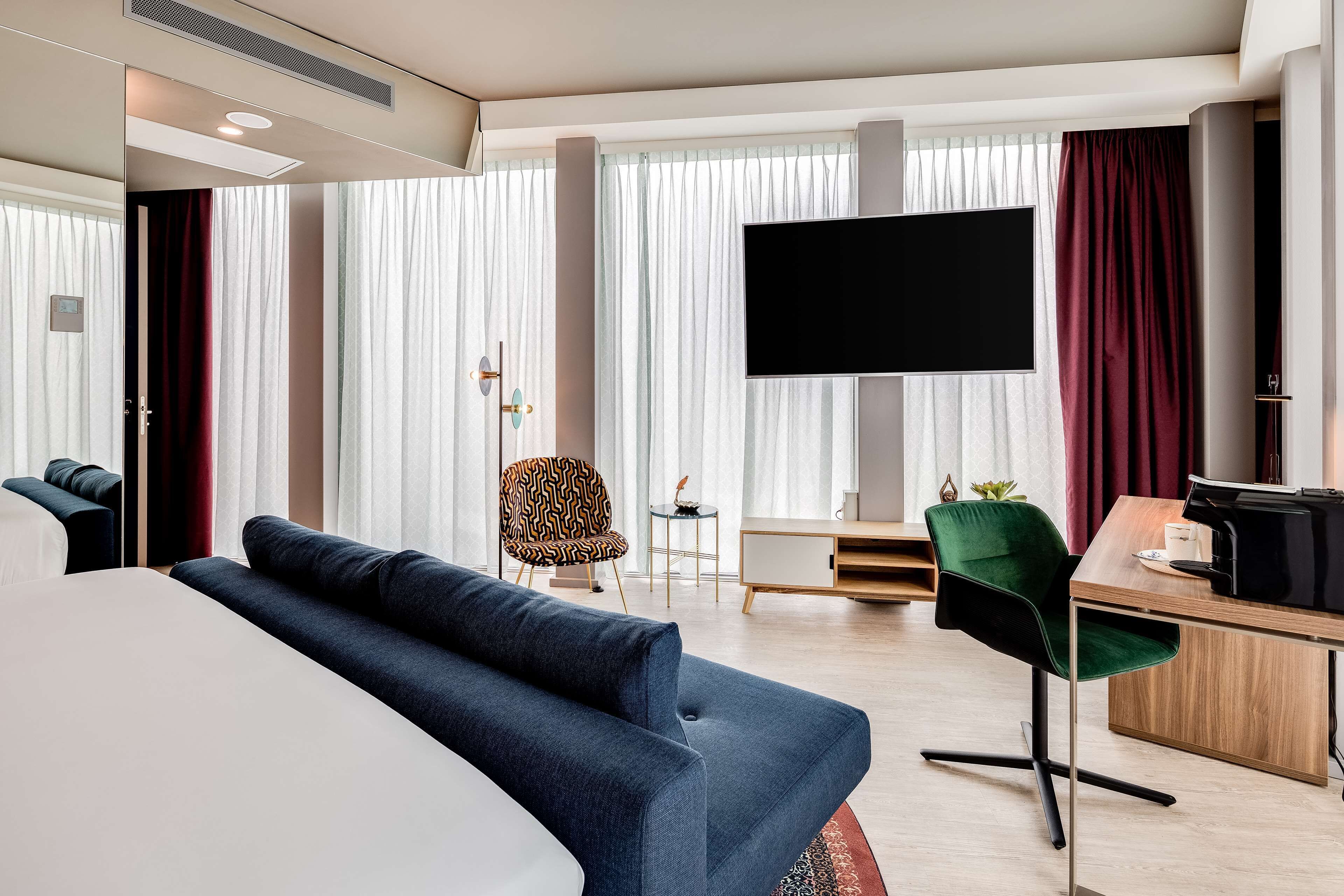 A modern hotel room with a large bed, a blue sofa, a desk with a computer, and a TV mounted on the wall.