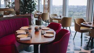 Un restaurante con una mesa alta dispuesta para comer, rodeado de sillas y grandes ventanales que ofrecen vistas a la ciudad.