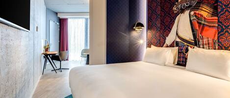 A hotel room with a large bed, a desk with a chair, a wall-mounted lamp, and a patterned tapestry.
