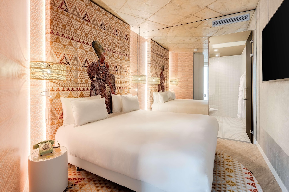 Habitación de hotel moderna con una cama grande, un diseño decorativo en la pared, una mesita con un teléfono y una zona de ducha.