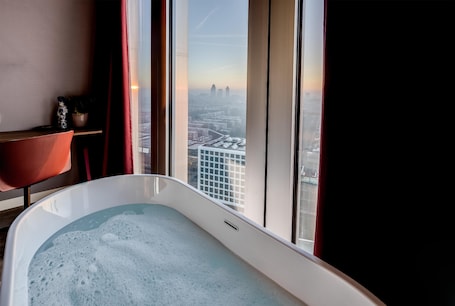 A modern bathroom with a bathtub, a red chair, and a view of a cityscape through large windows.. nhow Amsterdam RAI