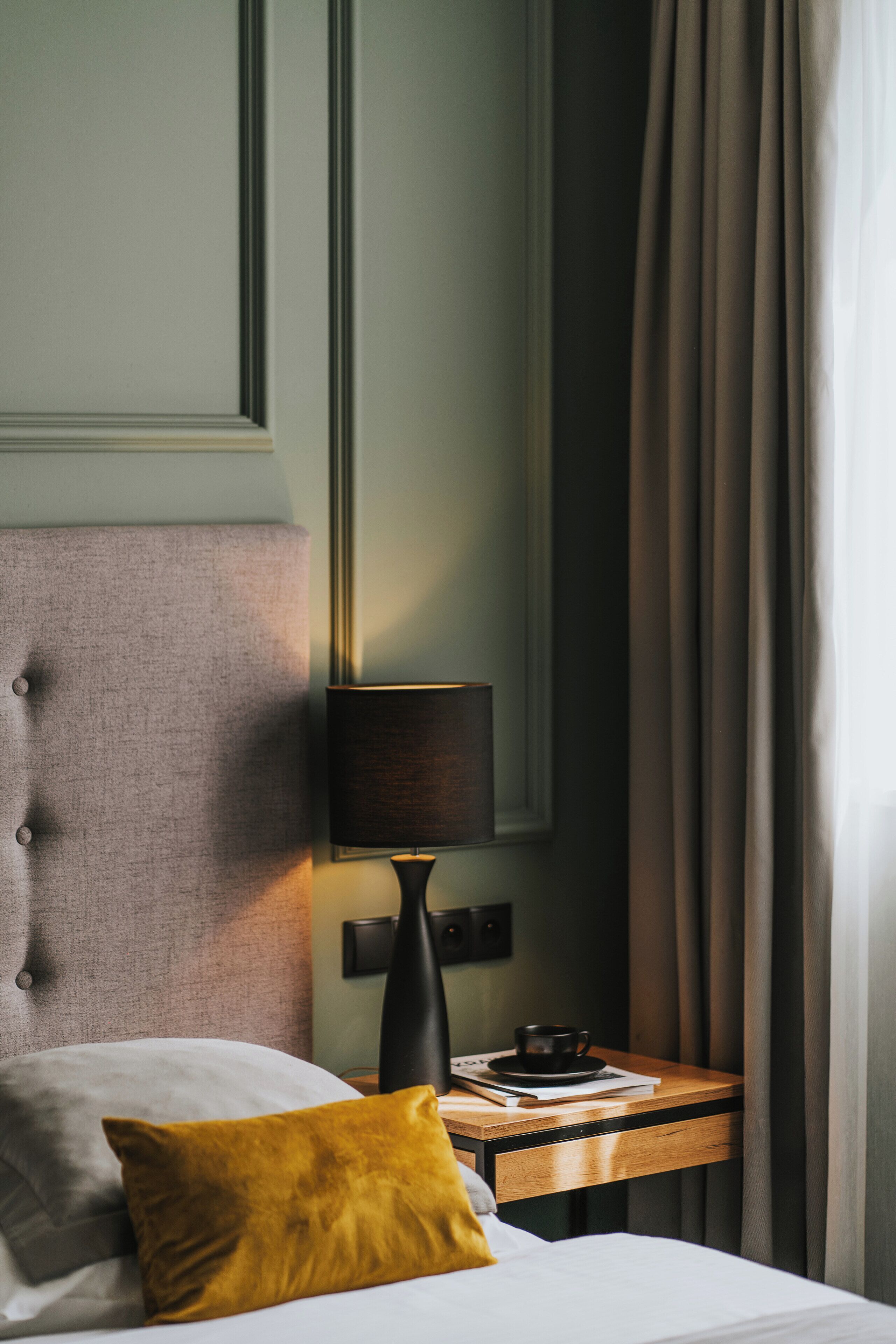 A neatly made bed with a white comforter and a mustard-colored pillow, a wooden nightstand with a lamp, a cup, and a book, and a large mirror on the wall.