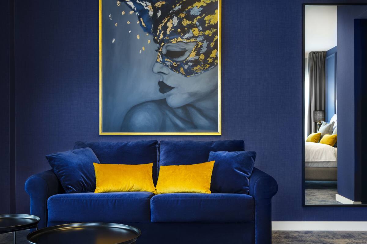 A modern room with a blue sofa, a framed artwork, and a mirror reflecting a bed with yellow pillows.
