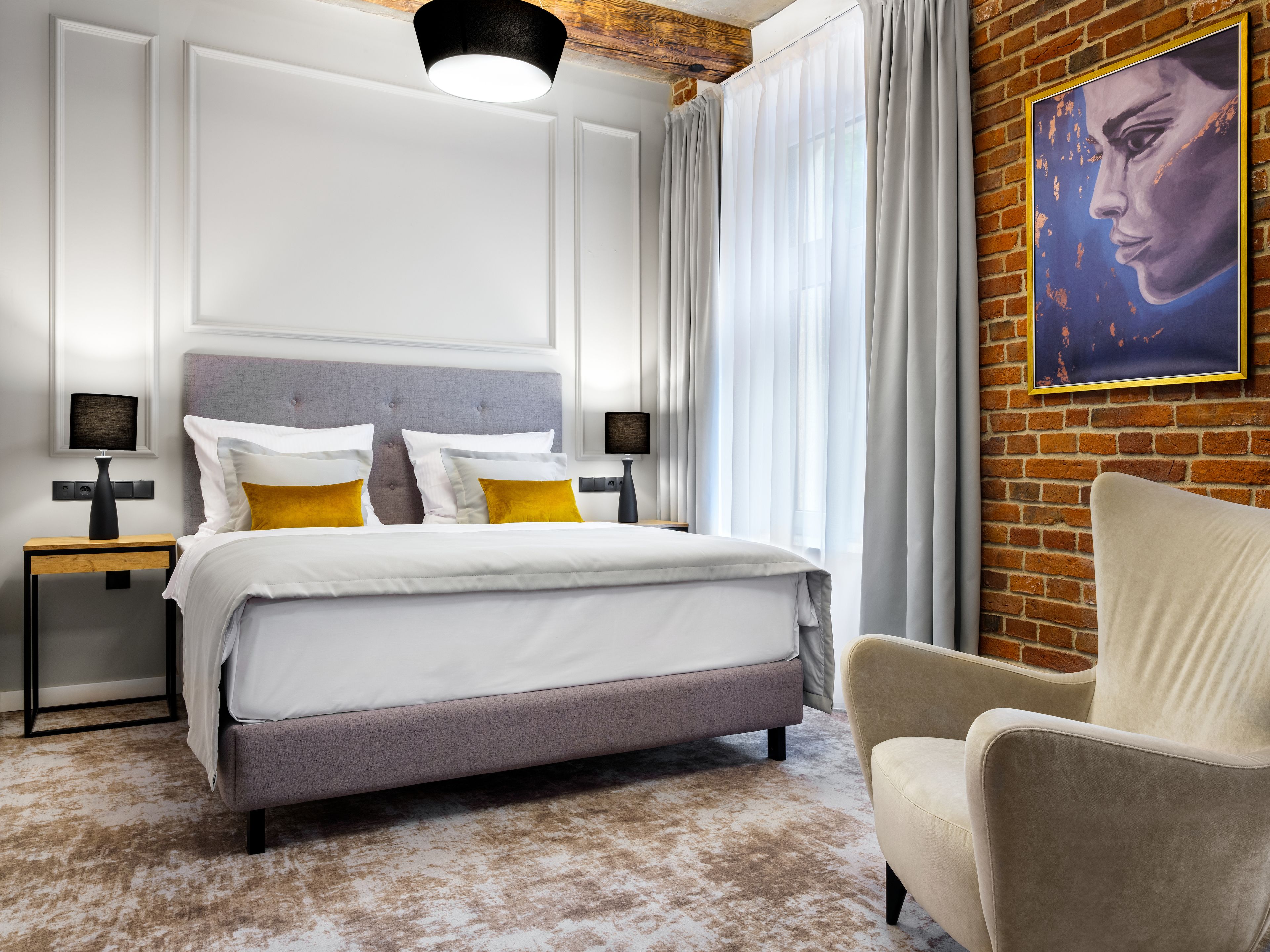 Un dormitorio moderno con una cama grande, dos mesitas de noche, una silla y una pared de ladrillo con un cuadro.