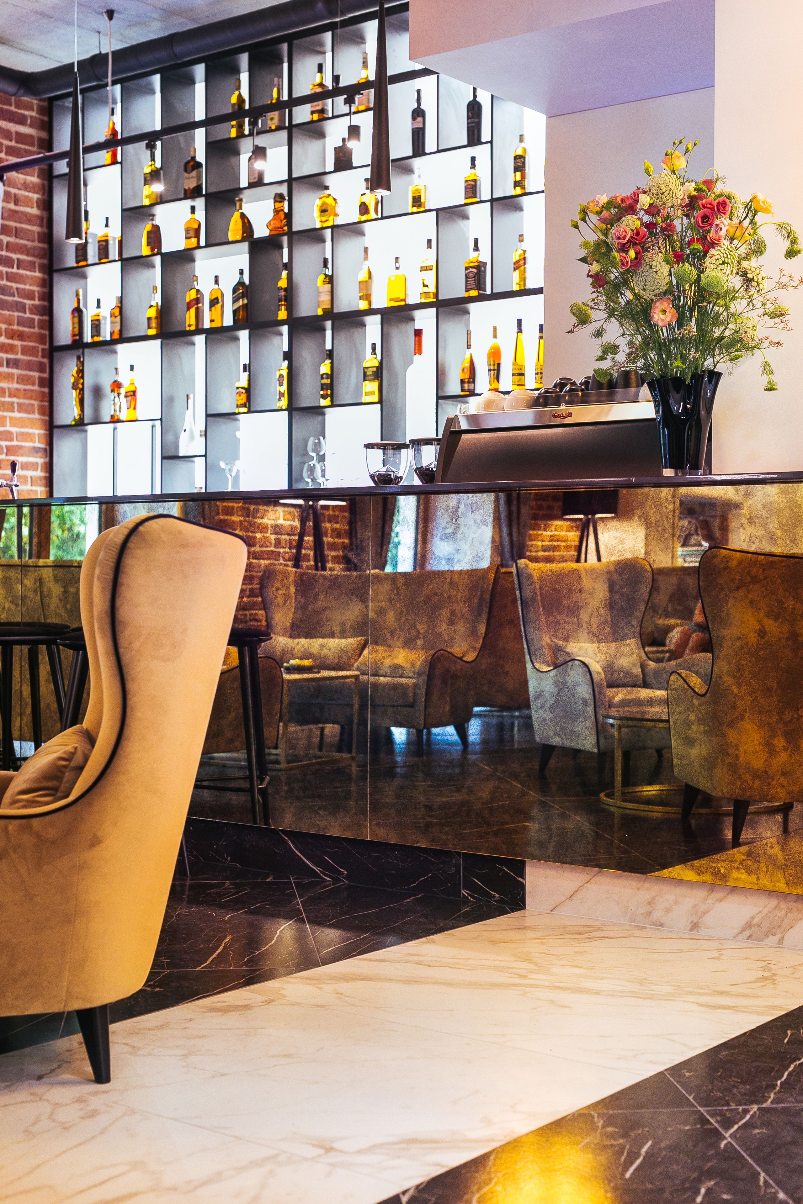 A bar area with a marble floor, a brick wall, and a shelf with various bottles. There are several chairs and a vase with flowers.