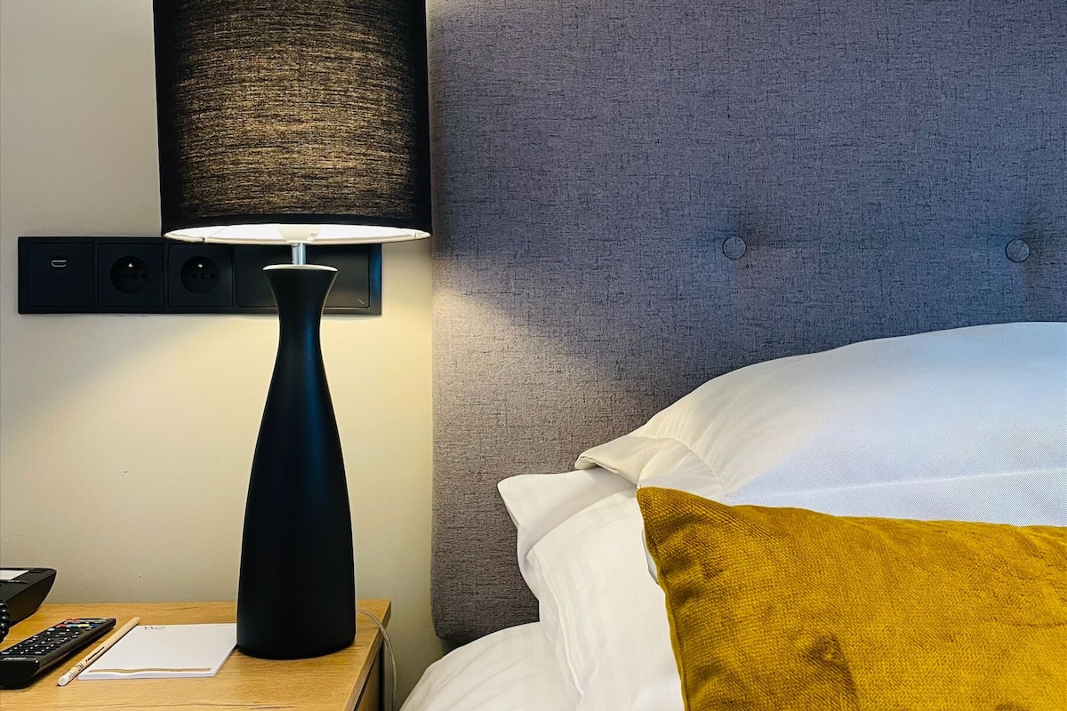 A neatly made bed with white and mustard pillows, a black lamp with a textured shade, and a wooden nightstand with a remote control and a notepad.