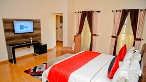 A hotel room with a bed, a TV mounted on a wooden panel, a desk, and a chair. - Aden 360 Apartments By Intra (Lagos)
