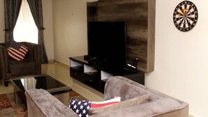 A living room with a flat-screen TV mounted on a wooden wall, a grey sofa with an American flag pillow, and a dartboard on the wall. - Aden 360 Apartments By Intra (Lagos)