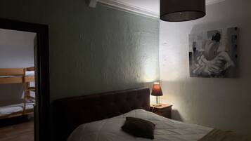 A bedroom with a single bed, a nightstand with a lamp, a painting on the wall, and a wooden door leading to another room.