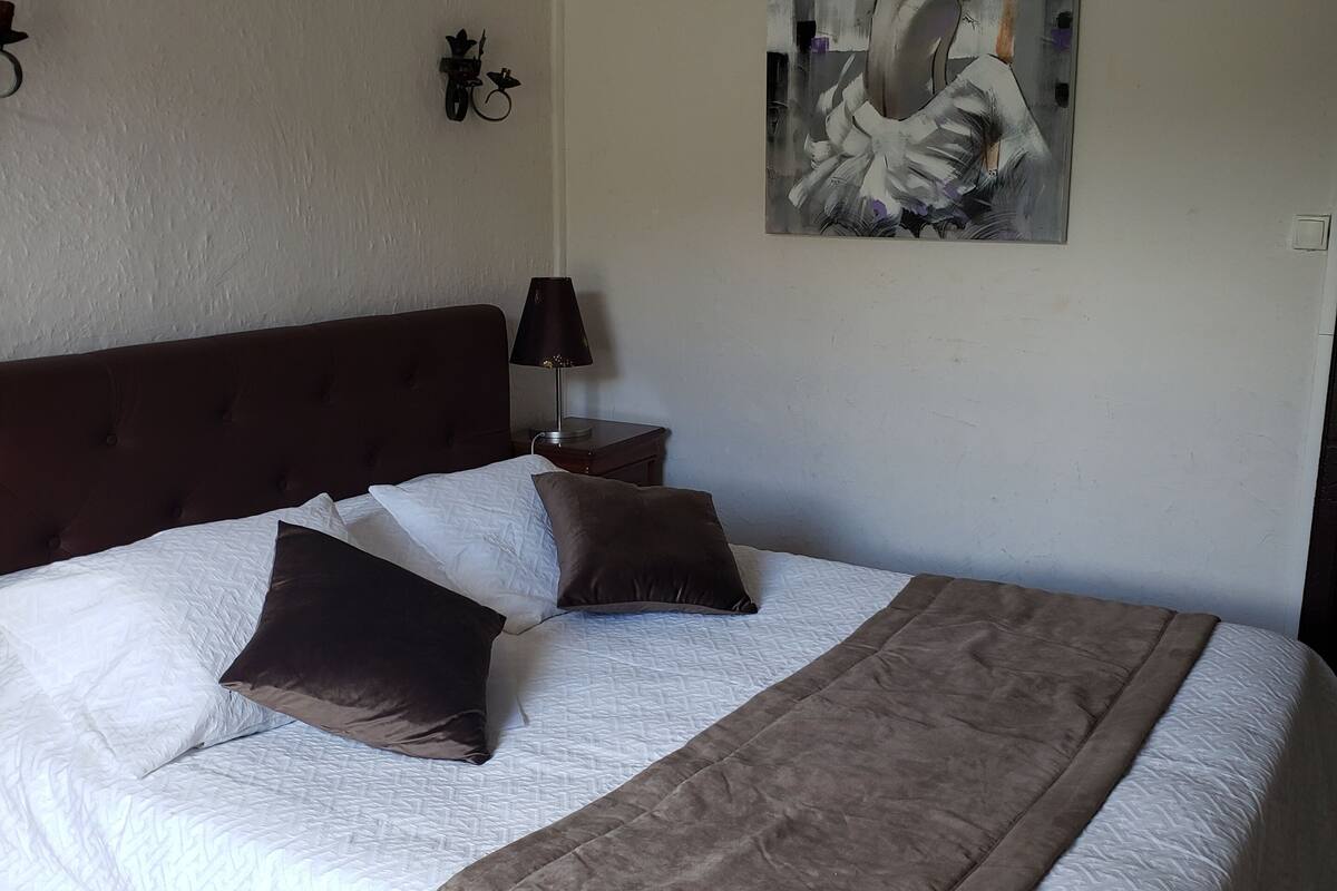 Een net opgemaakt bed met een bruin hoofdeinde, een witte quilt en meerdere kussens. Boven het bed hangt een schilderij aan de muur.