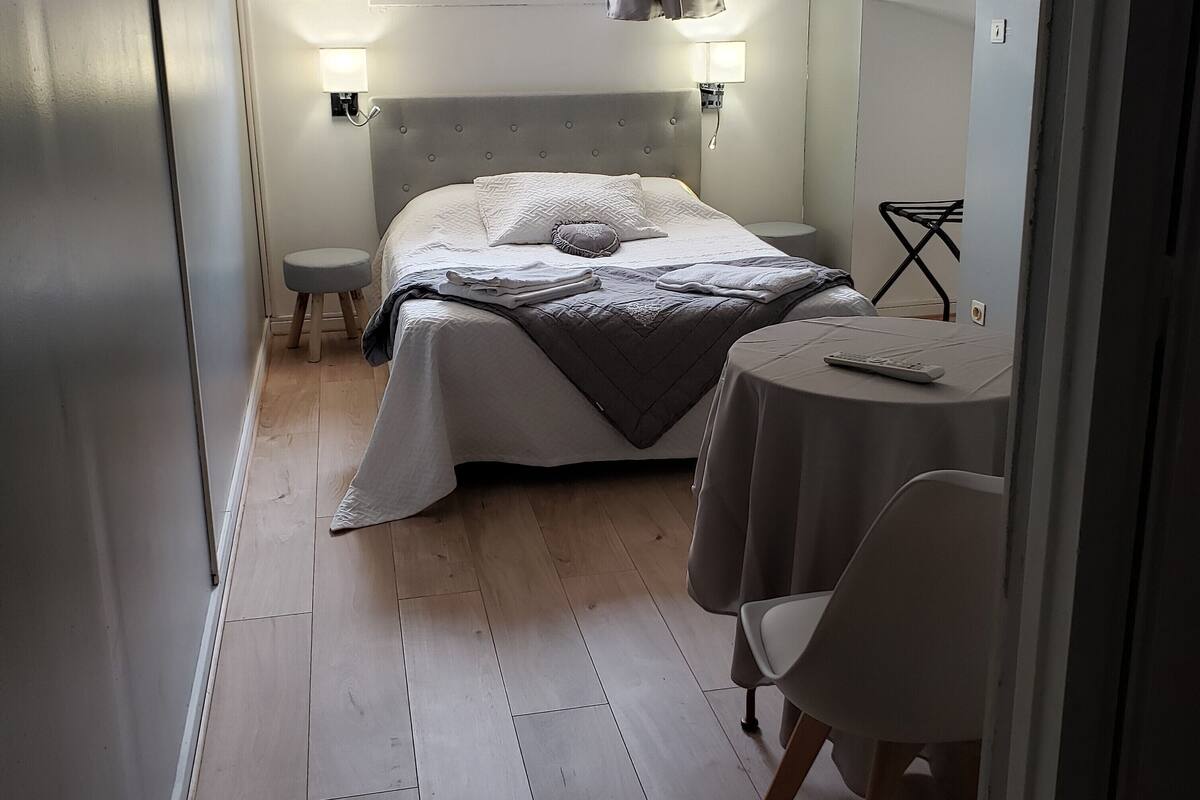 Een knus slaapkamer met een bed, nachtkastjes, een klein krukje, een tafel met stoel, en een raam met uitzicht op bomen.
