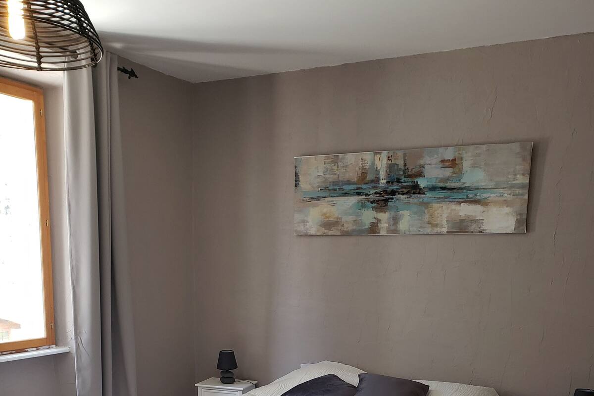 Een slaapkamer met een groot bed, een nachtkastje, een lamp, een schilderij aan de muur en een raam met gordijnen.