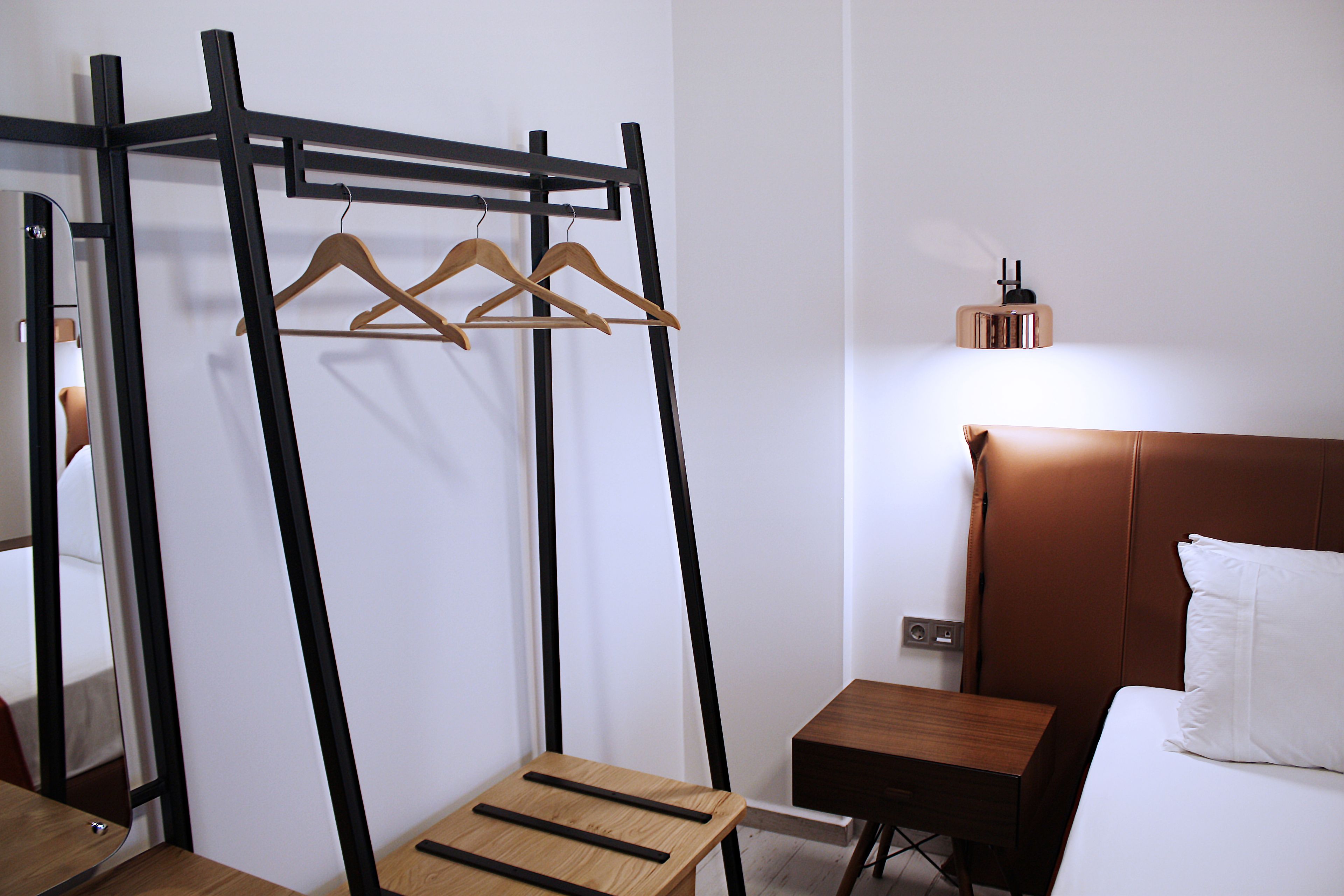 A hotel room with a bed, a bedside table, a wooden chair, a coat rack, and a mirror.