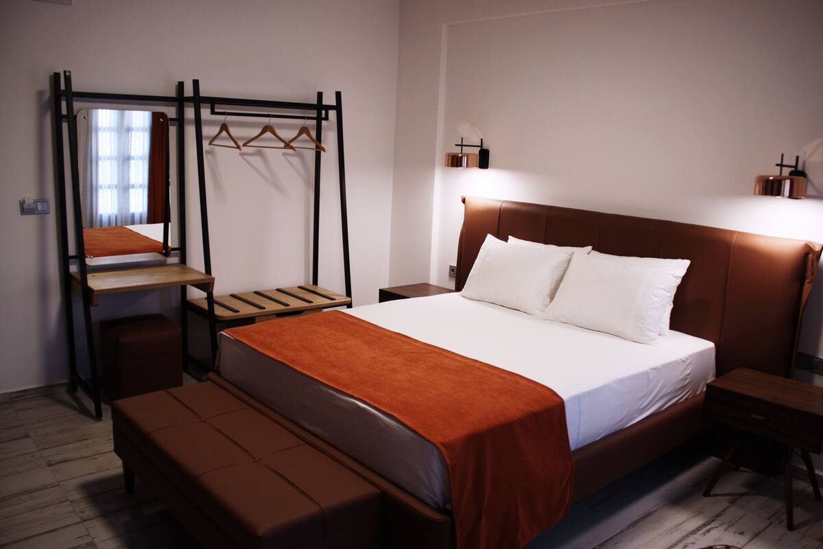 A bedroom with a bed, a bench, a hanging clothes rack, and a bedside table.