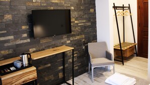 A modern room with a stone wall, a flat-screen TV, a wooden side table with a kettle, a patterned chair, a wooden coat rack, and a door. - Pintura Urla Butik Otel & Sanat Galerisi (Urla)