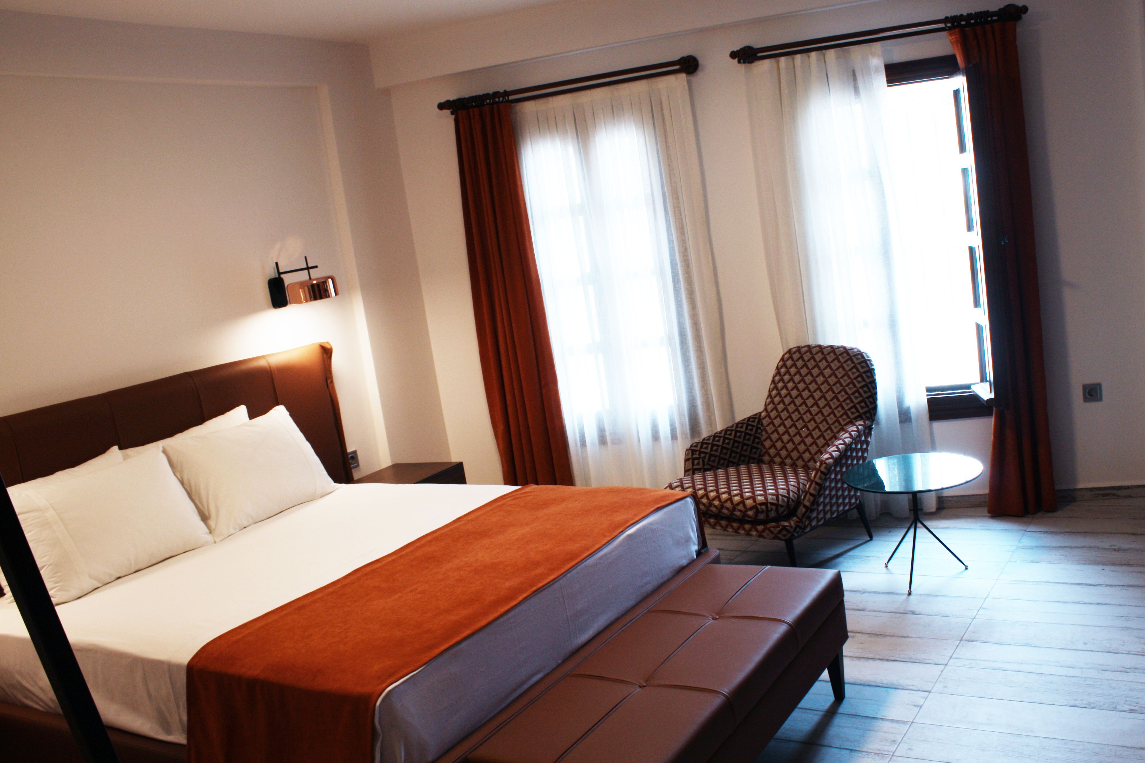 A hotel room with a large bed, a small round table, a patterned armchair, and curtains on the windows.