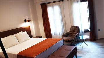A hotel room with a large bed, a small round table, a patterned armchair, and curtains on the windows.