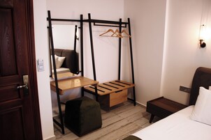 Habitación de hotel compacta con cama, escritorio pequeño, banco de madera, perchero y mesita de noche.