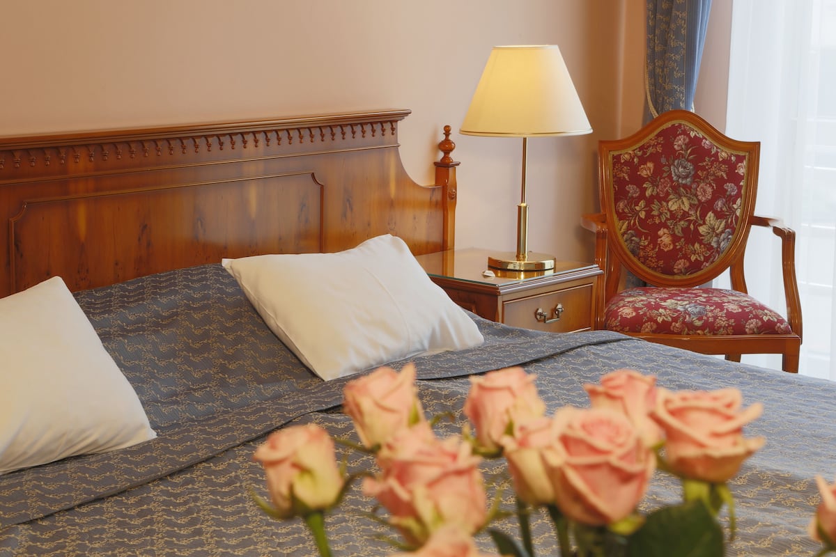 A neatly made bed with a floral-patterned bedspread, a wooden headboard, a bedside table with a lamp, and a floral-patterned chair.