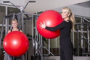 Zwei Personen trainieren mit großen roten Fitnessbällen in einem Fitnessstudio mit Metallgeräten und Fenstern.