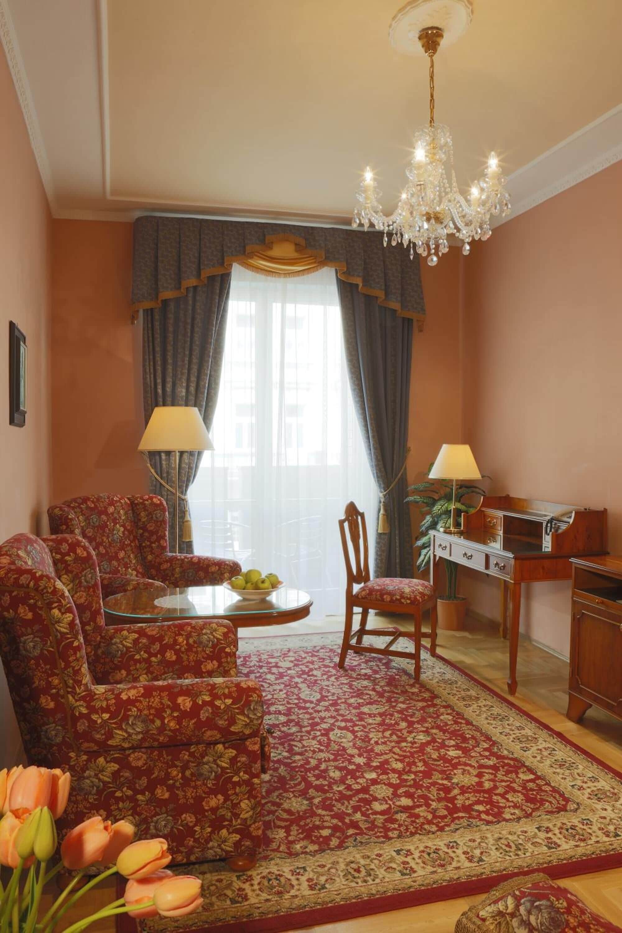 Una habitación con candelabro, sillones tapizados con motivos florales, un escritorio con lámpara y una alfombra roja estampada.