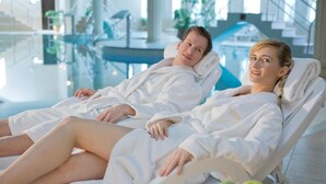 Two individuals in white bathrobes lying on reclining chairs in a well-lit indoor setting with a glass floor and a staircase in the background. - Hotel Bristol (Karlovy Vary)