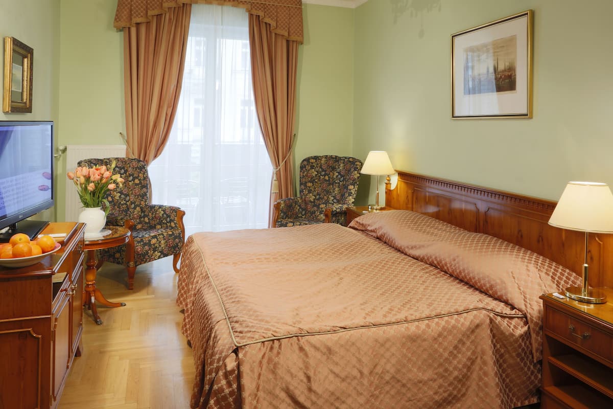 A hotel room with a bed, two chairs, a desk with a computer, and a window with curtains.