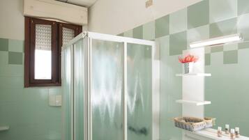 A bathroom with a glass shower enclosure, a sink, a mirror, and a window with blinds.