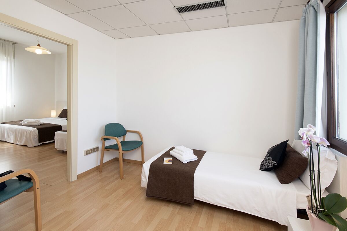 Une chambre d’hôtel avec un lit simple, une chaise, une petite table et une fenêtre avec des rideaux.