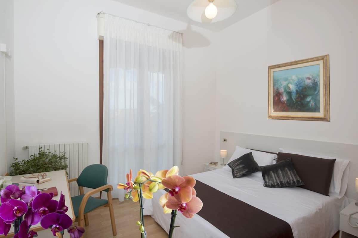 A hotel room with a bed, a desk with a chair, a window with sheer curtains, and a painting on the wall.