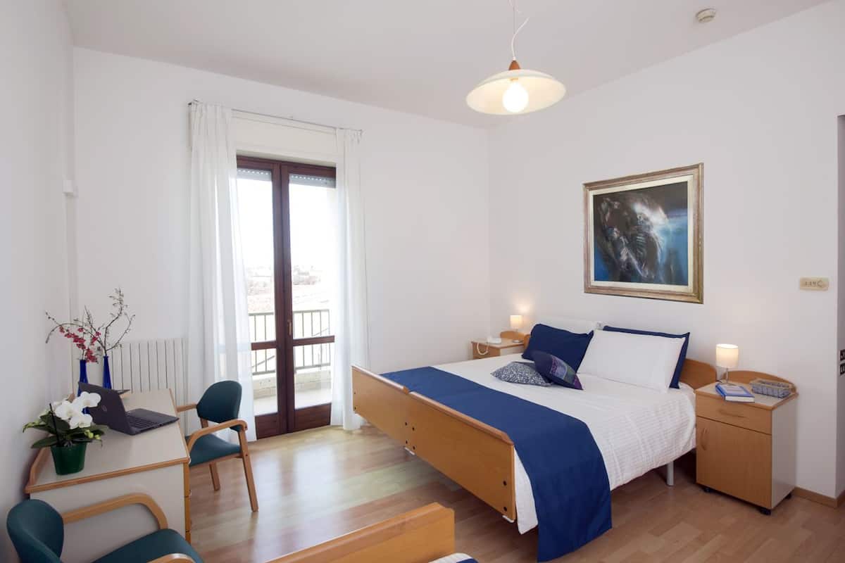A bedroom with a bed, bedside tables, a desk with a chair, a potted plant, and a window with curtains.