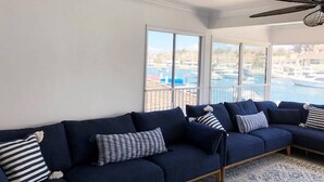 Un salon avec un canapé bleu foncé, un tapis à motifs et une vue sur une marina, visible à travers de grandes fenêtres.