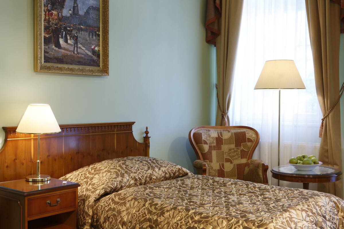 Een hotelkamer met een bed, nachtkastje, stoel, klein tafeltje met een schaal fruit en een schilderij aan de muur.