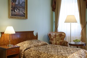 Een hotelkamer met een bed, nachtkastje, stoel, klein tafeltje met een schaal fruit en een schilderij aan de muur.