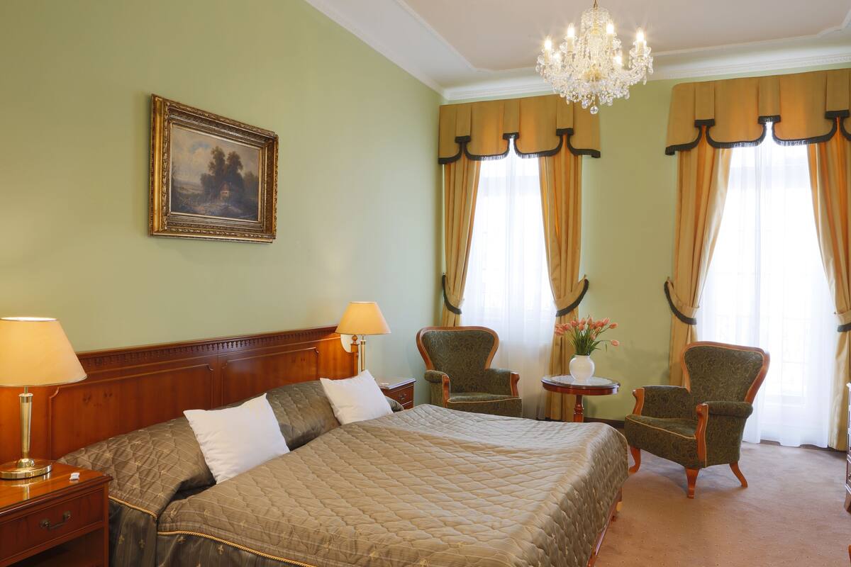 Een hotelkamer met een groot bed, twee stoelen, een tafeltje met een vaas en een kroonluchter.