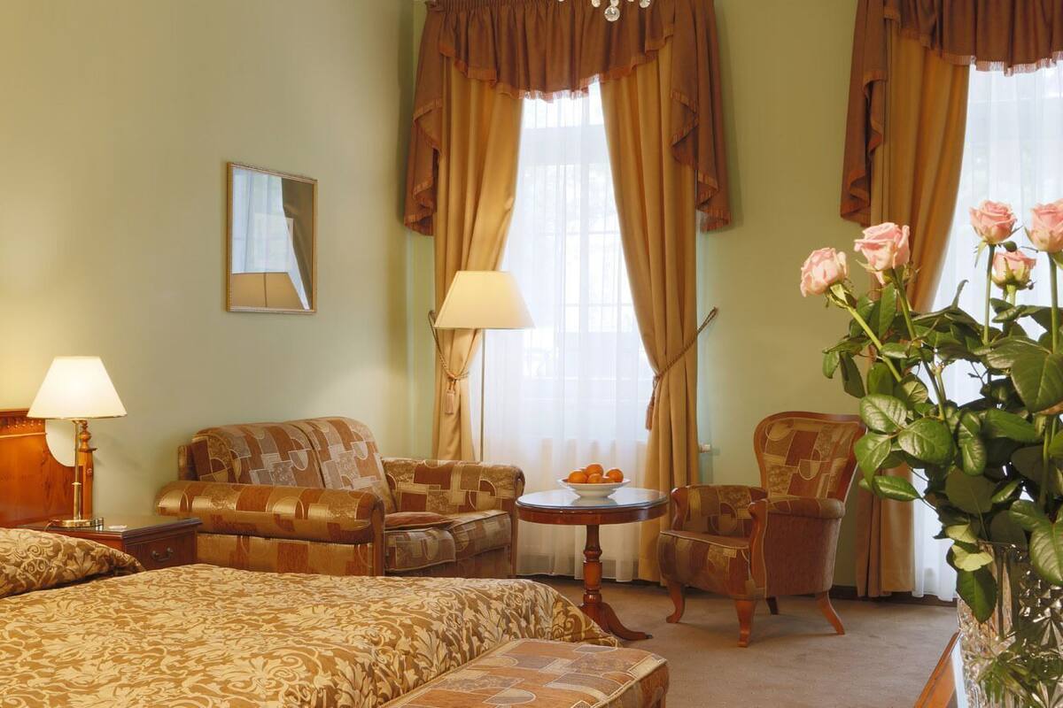Een hotelkamer met een bed, twee fauteuils, een tafeltje, een lamp, een spiegel en een bloemenvaas.